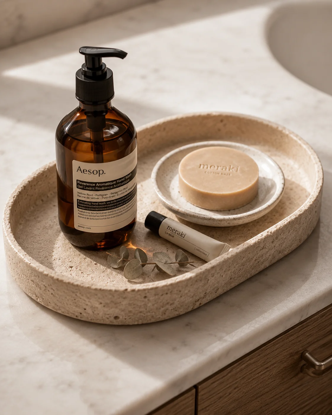 A warm travertine stone oval tray on a marble bathroom countertop holding an Aesop amber glass soap dispenser, a ceramic soap dish with a bar of soap, and a small eucalyptus sprig