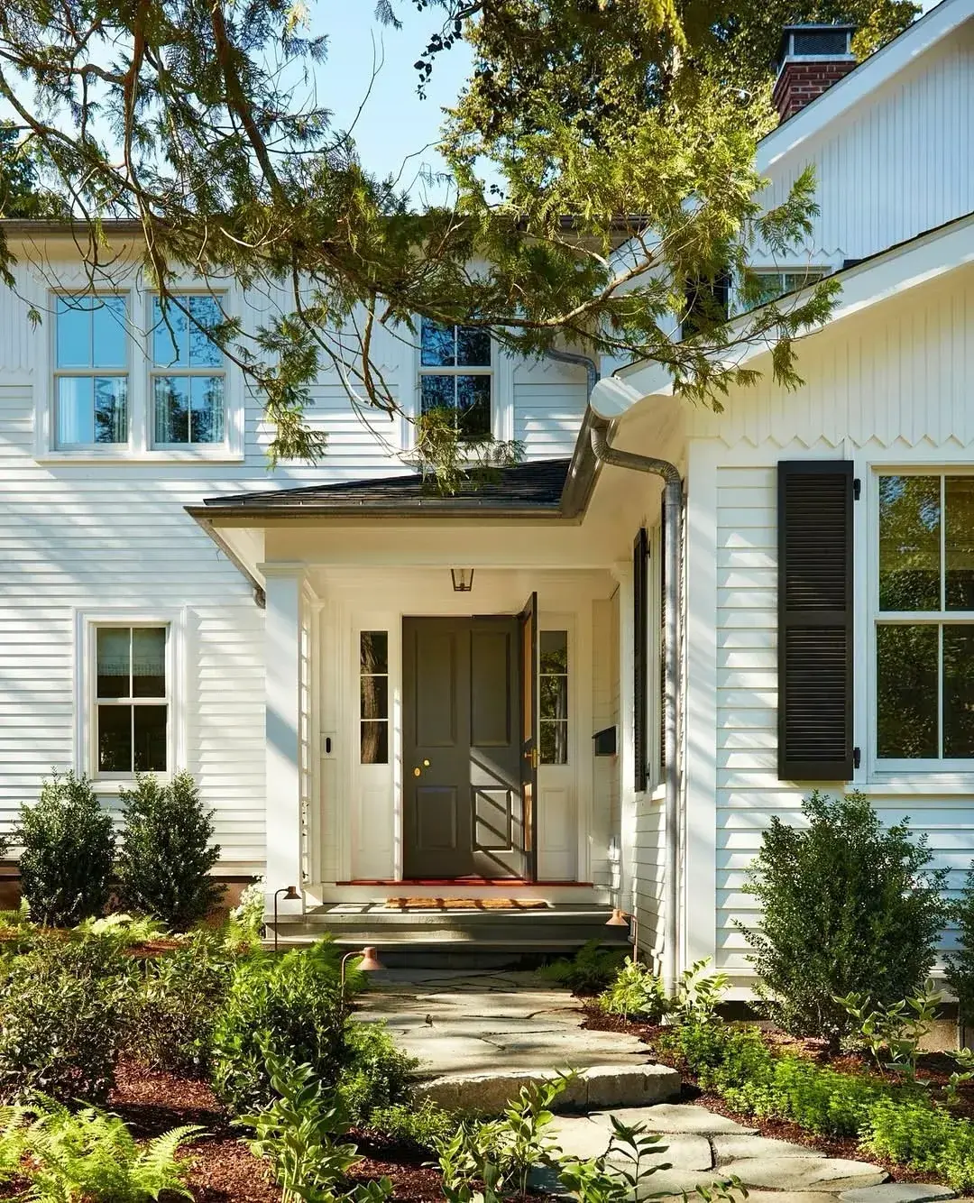 White clapboard house in full sun with a dark door ajar, black shutters and a stone path