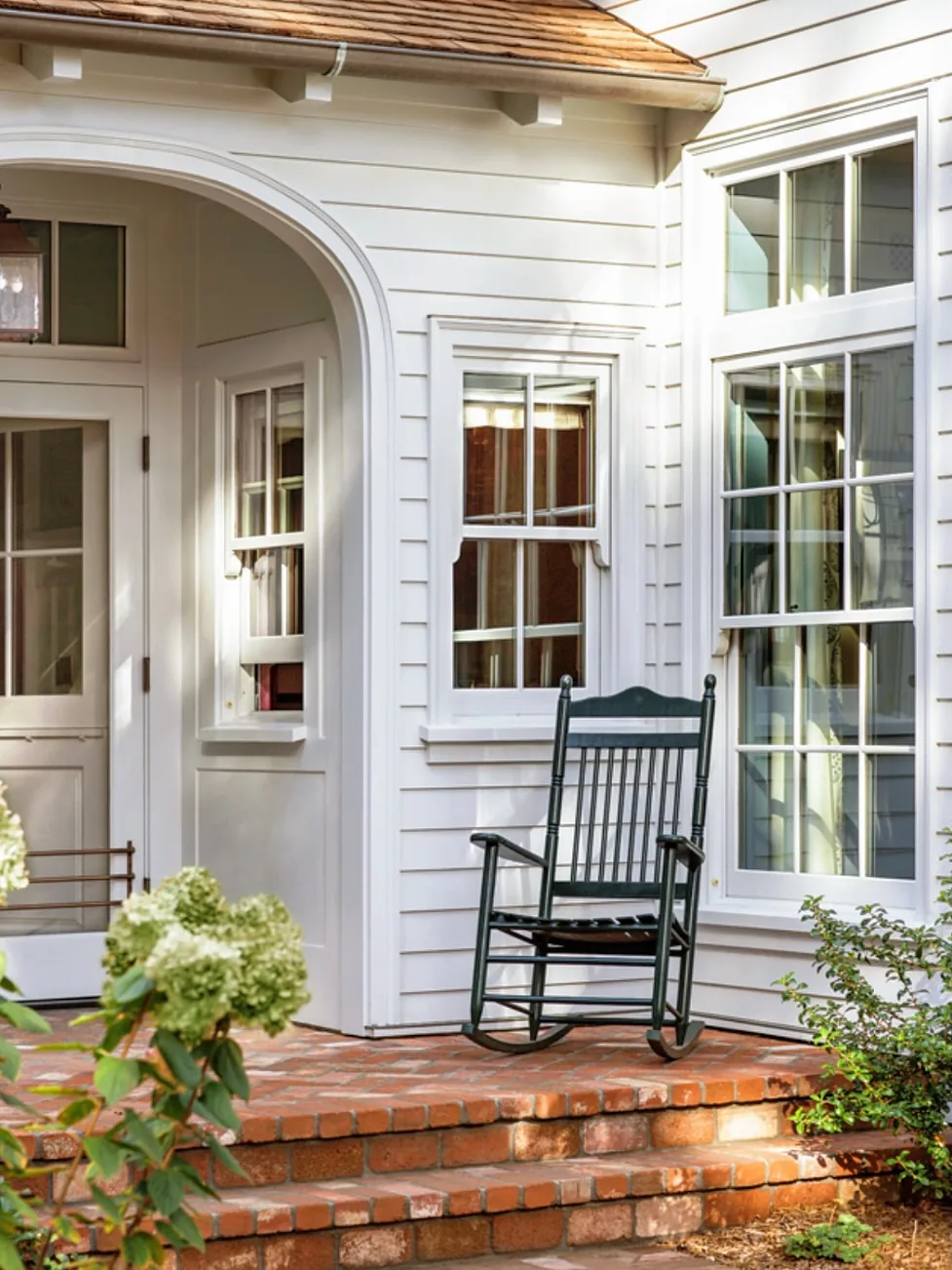 White cottage porch corner with a black rocking chair on brick steps beside tall windows