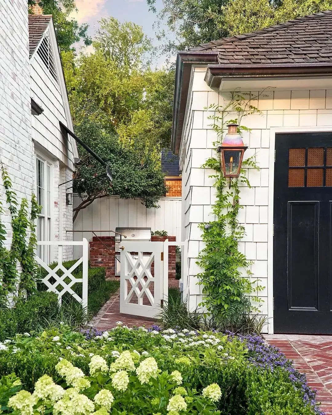 White shingle cottage with a black panelled door with amber glazing, a copper lantern and a white gate