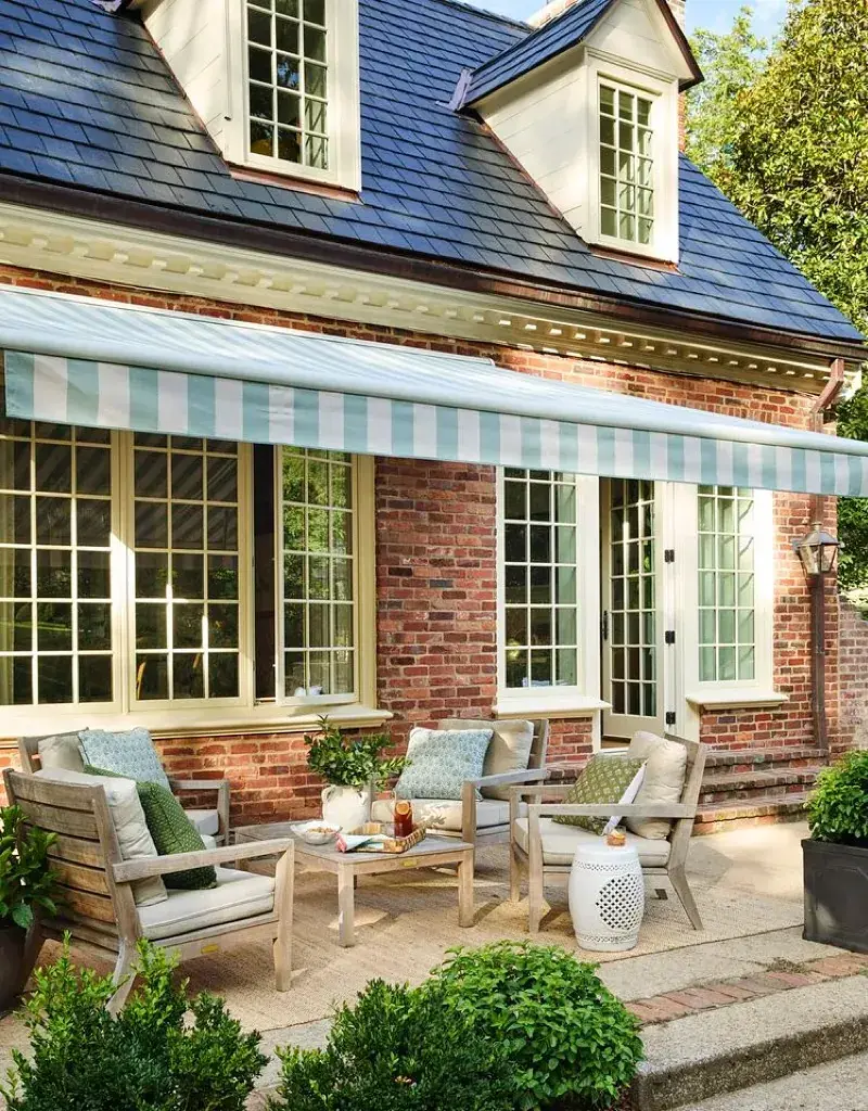 Red brick house with striped awning, teak chairs and rug over patio