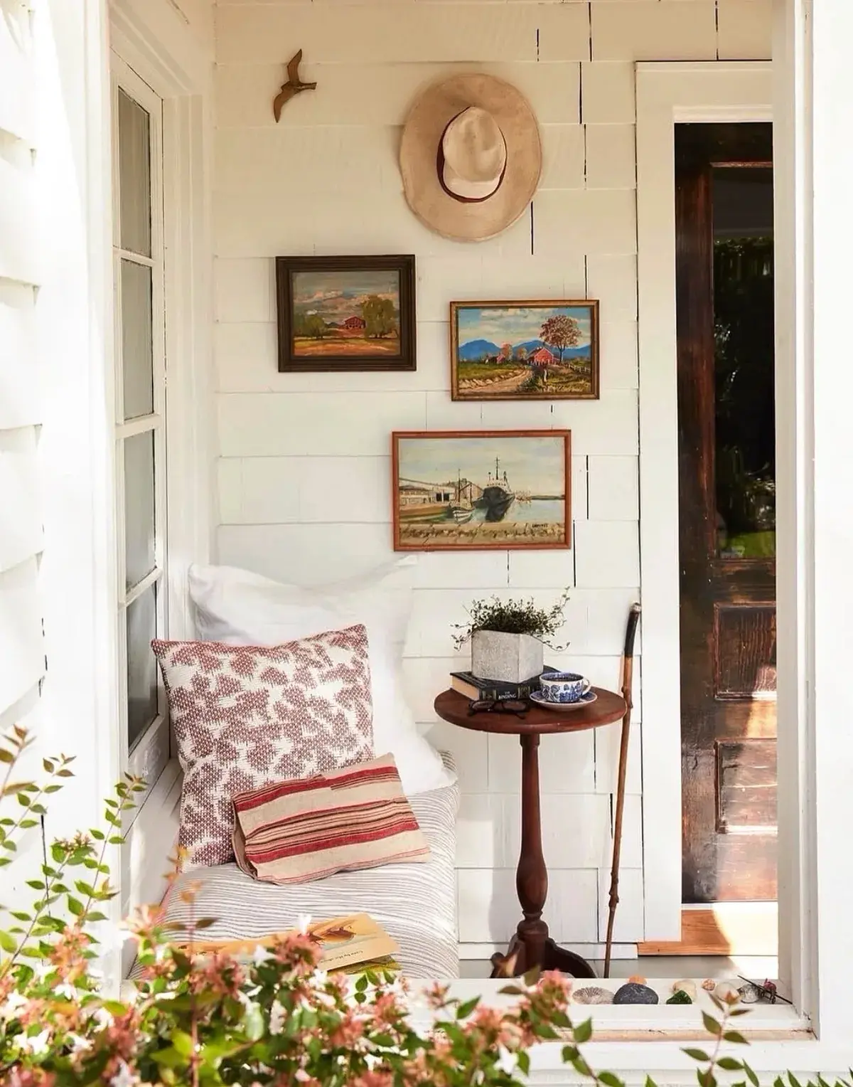 Small enclosed porch corner with a striped daybed, cushions, a hat and three small paintings on white shingle wall