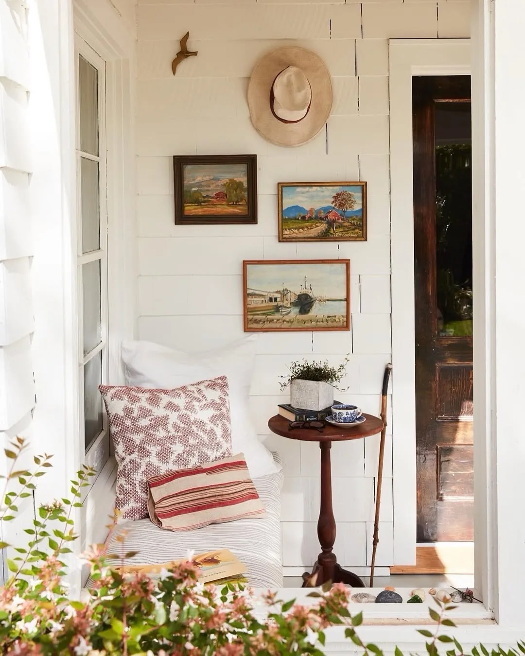 Porch wall with a straw hat, three small framed paintings, and a small round table beside the daybed