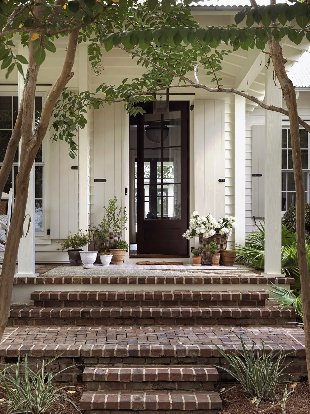 White farmhouse entrance with wide brick steps, a dark panelled door, and baskets of white flowers by the threshold