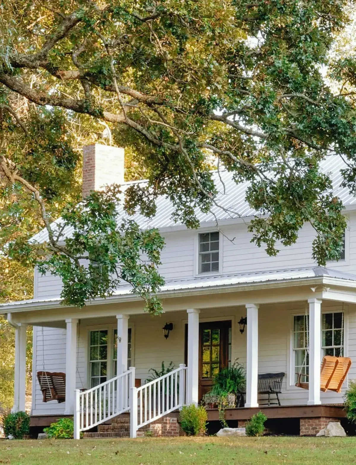Large white farmhouse with a wraparound porch, white rails, hanging swings and a dark stained deck