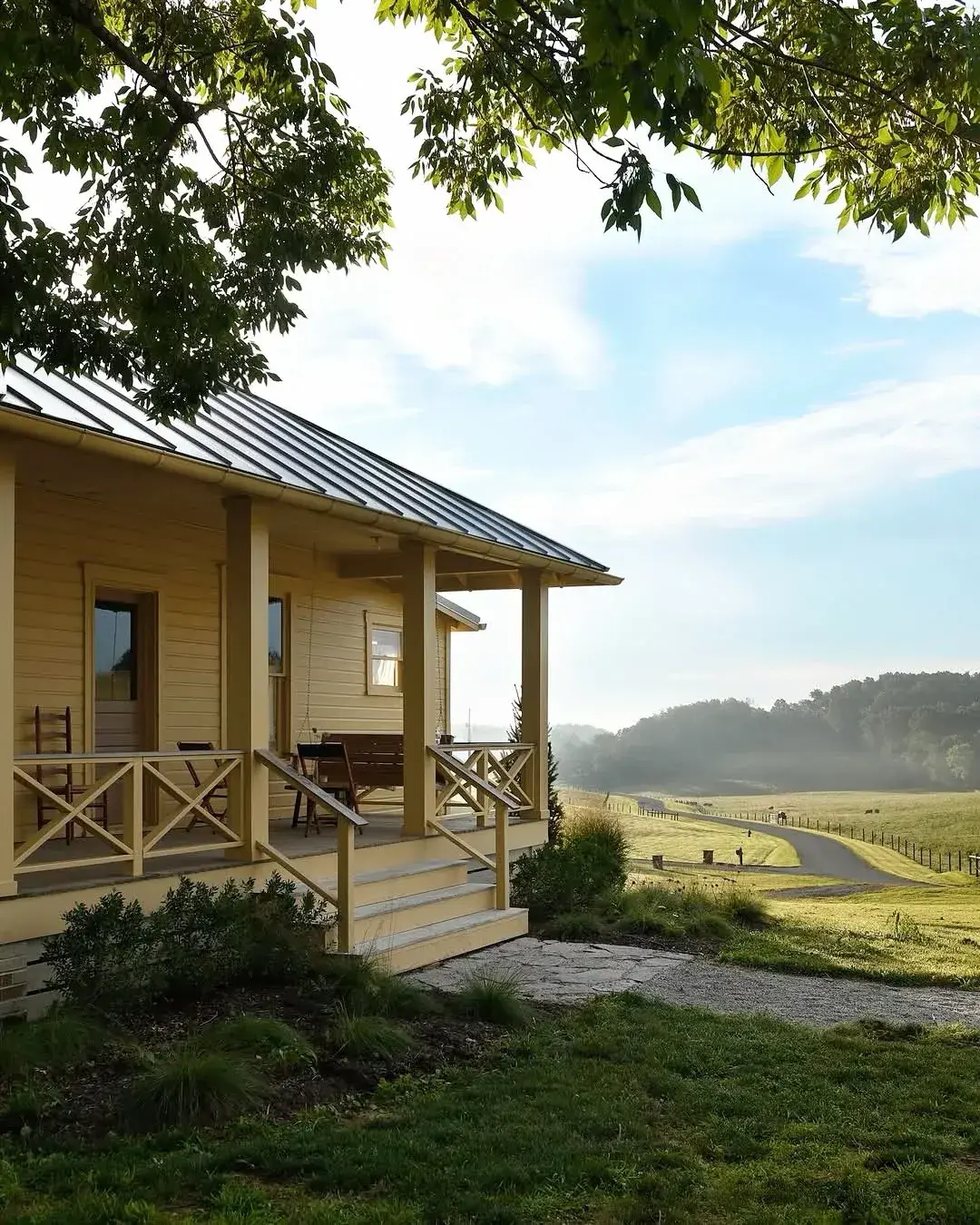 Pale yellow house with a deep porch, cross braced rails, a metal roof and steps to a meadow beyond
