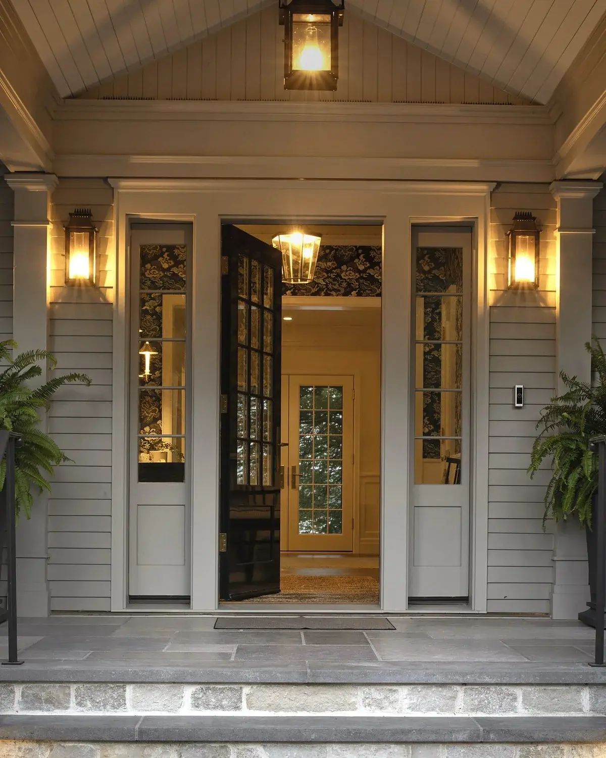 Grey weatherboard entry at dusk with glazed door, sidelights and lit lanterns