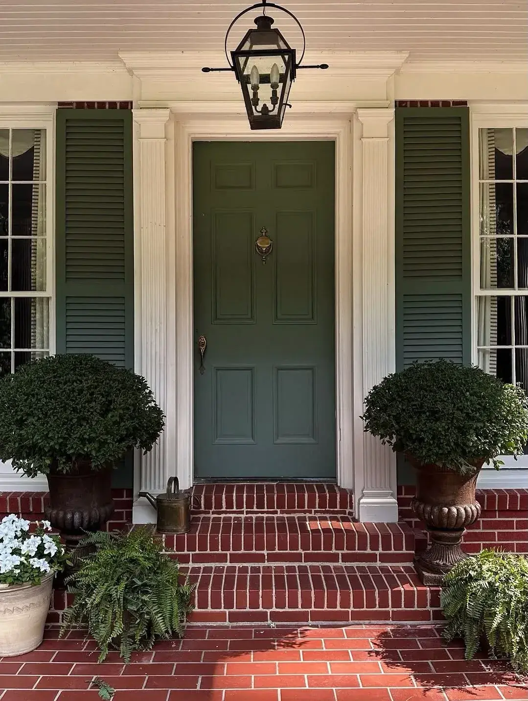 Olive green door with matching shutters on red brick porch with boxwood urns