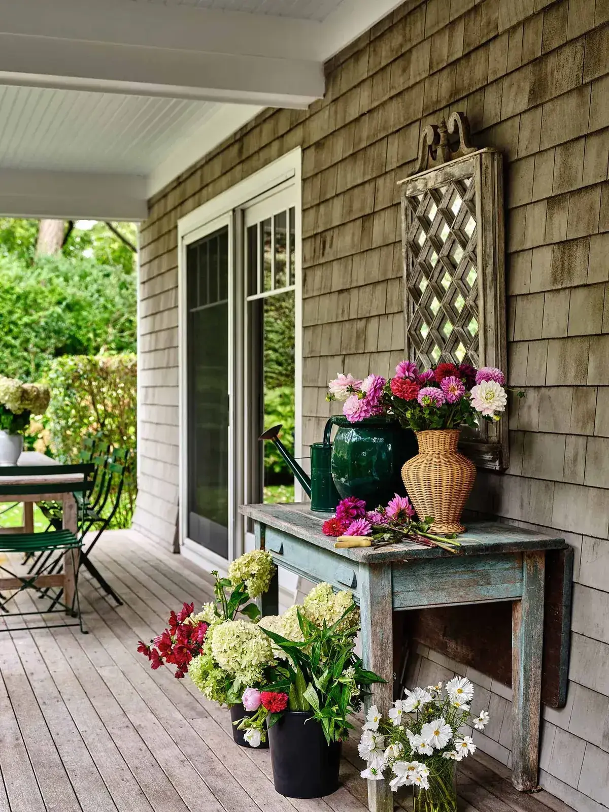 Weathered table on a shingle porch holding cut dahlias, pale hydrangeas in buckets, and a watering can