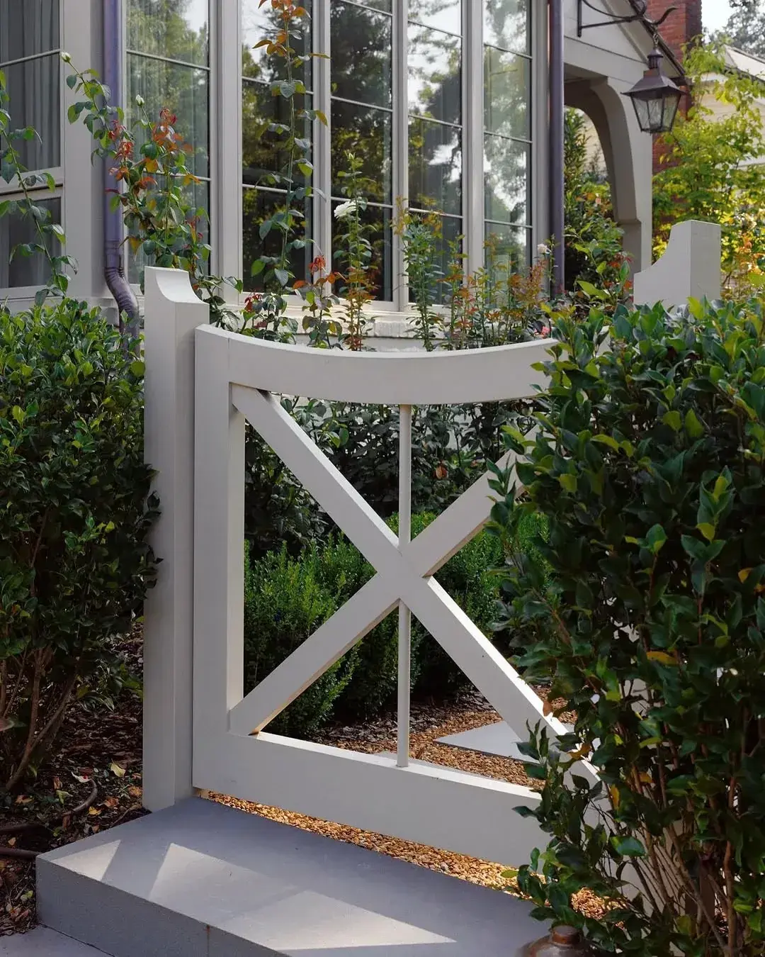 White crossbar gate between tall box hedges with roses and tall windows behind