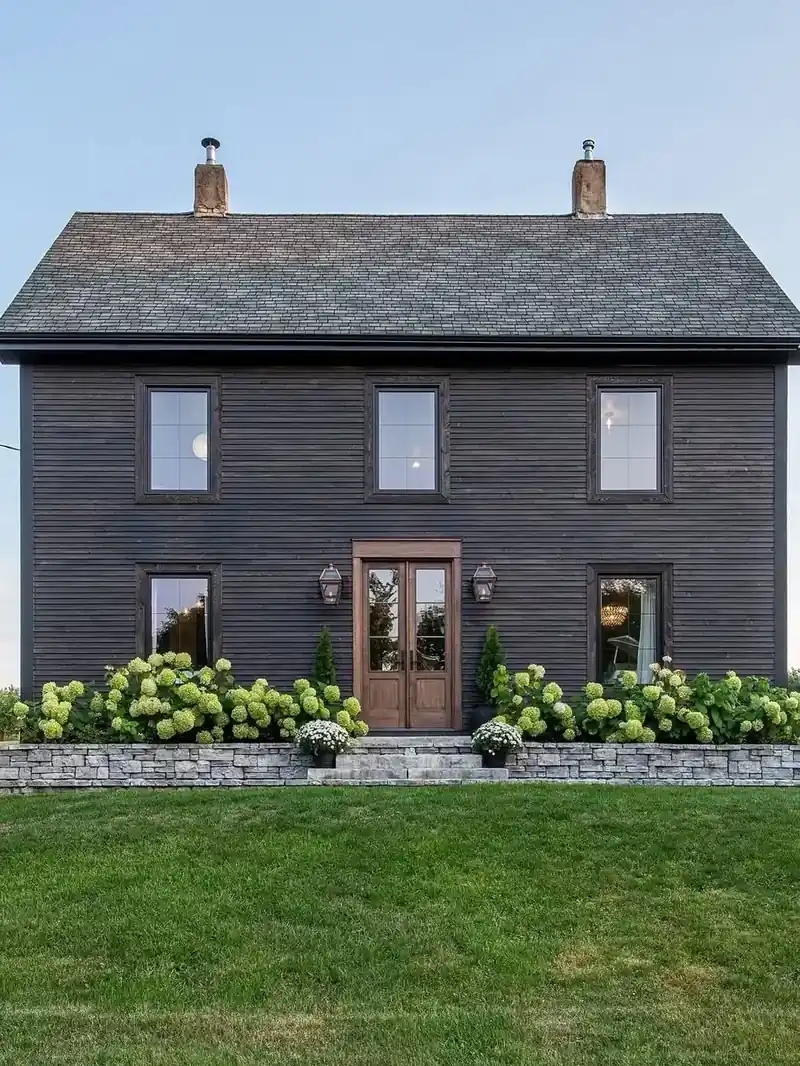 Black clapboard farmhouse with a timber door, a low stone wall and lime hydrangeas along the front