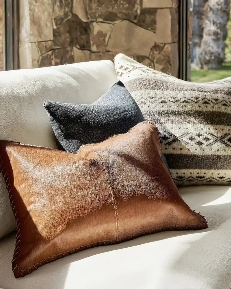 Cognac hair-on-hide, charcoal linen and cream knit cushions layered on a pale sofa against stone
