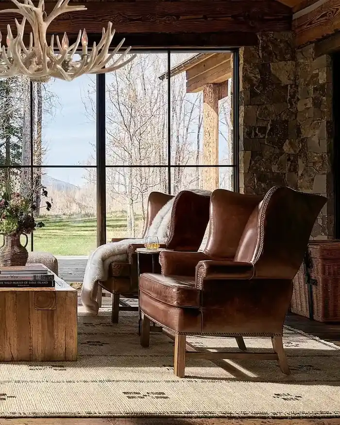 Two cognac leather wingback chairs with a shearling throw under a pale antler chandelier, facing a mountain view