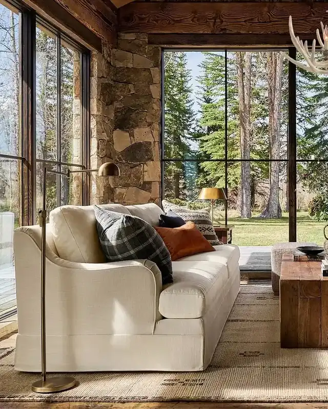 Cream slipcovered sofa with plaid and rust cushions beside a stone pier and tall black-framed windows onto a lawn