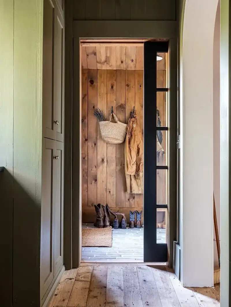 Mudroom with knotty pine walls, black iron hooks holding a straw basket and jacket, and boots on the floor