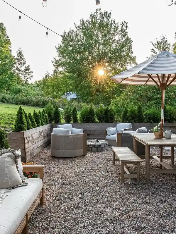 Gravel patio with wicker lounge chairs, a stone fire pit, a timber dining table and string lights overhead