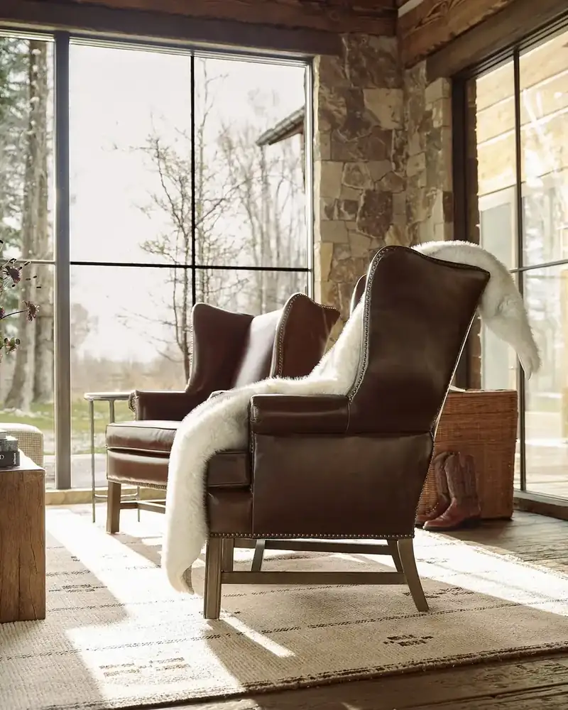 Two dark leather wingback chairs with cream shearling throws beside floor-to-ceiling windows onto winter trees