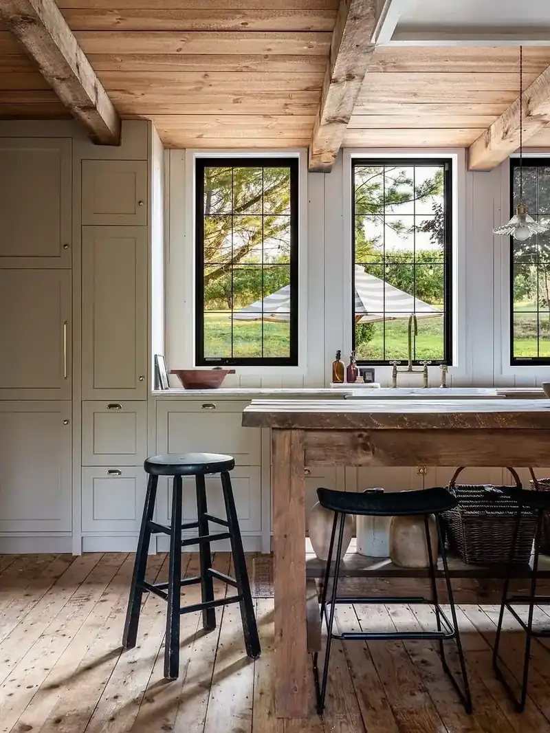 Rustic kitchen with a pale timber butcher-block island, black stools, grey-green cabinets and black-framed garden windows