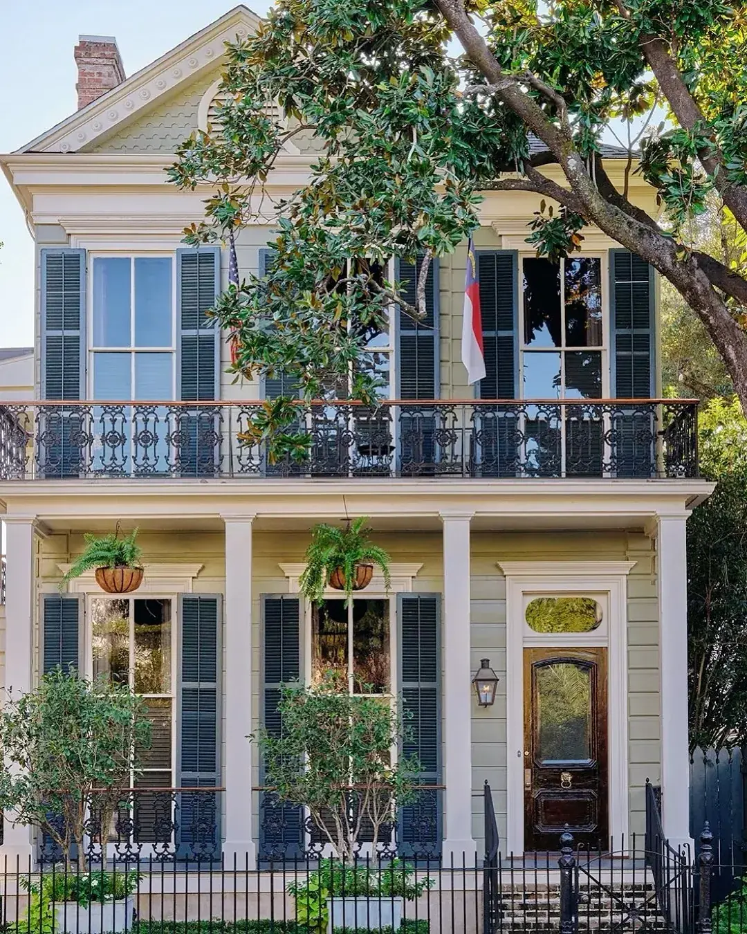 Pale yellow New Orleans house with dark blue-grey louvered shutters, wrought iron balcony and hanging ferns