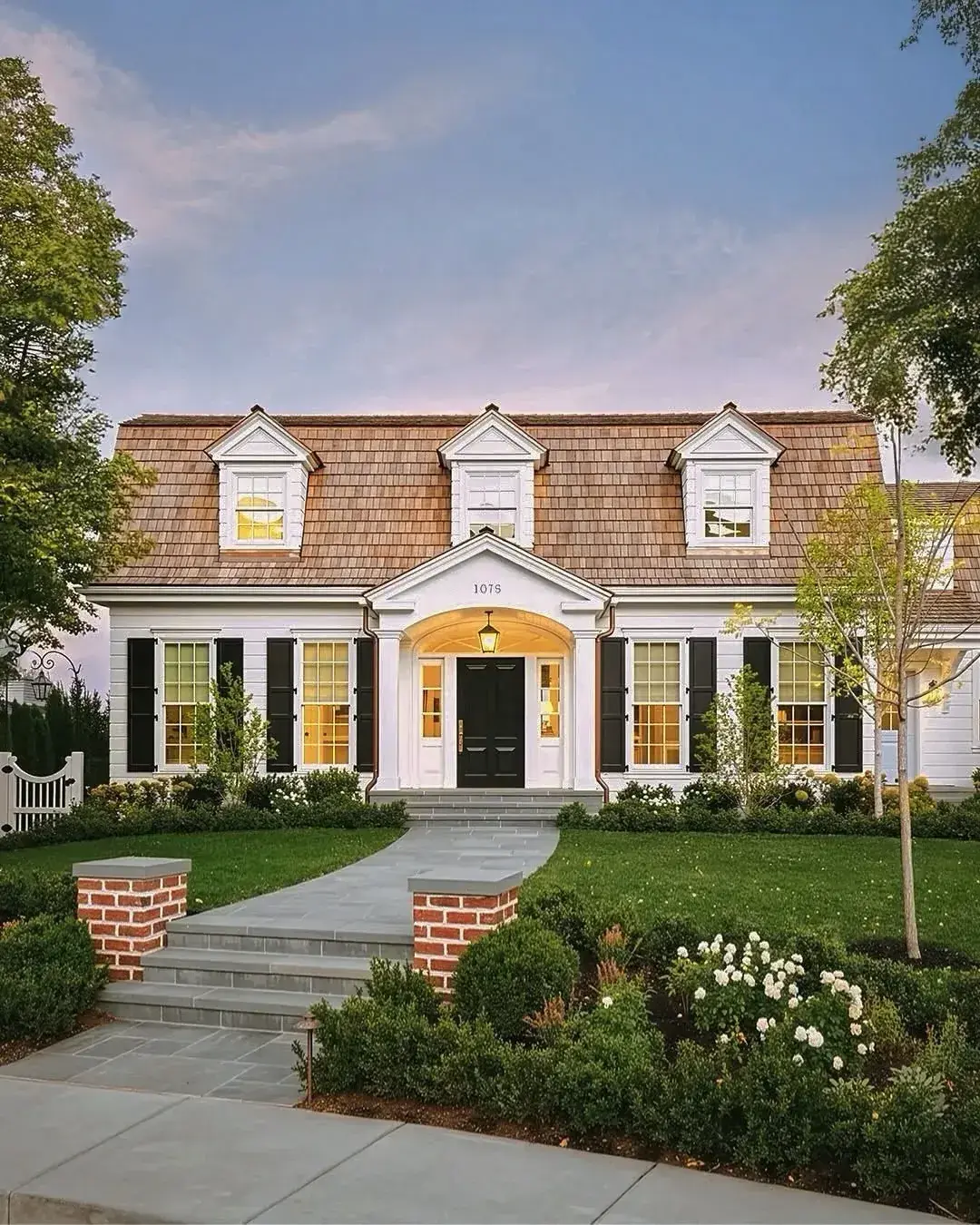 White clapboard Cape Cod with black louvered shutters, three dormers and an arched portico