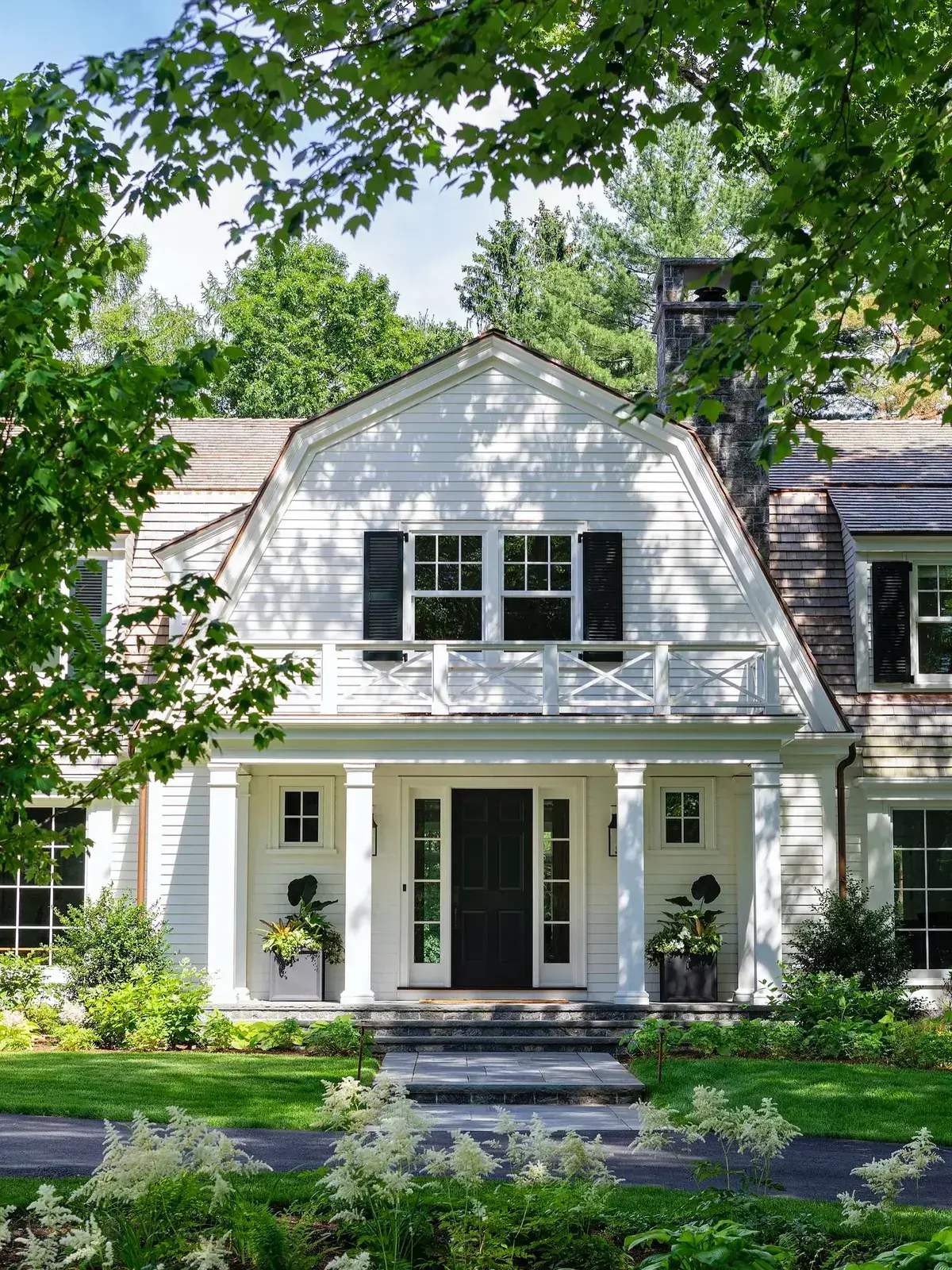 White gambrel house with black shutters on an upper paired window, X rail balcony and columned portico
