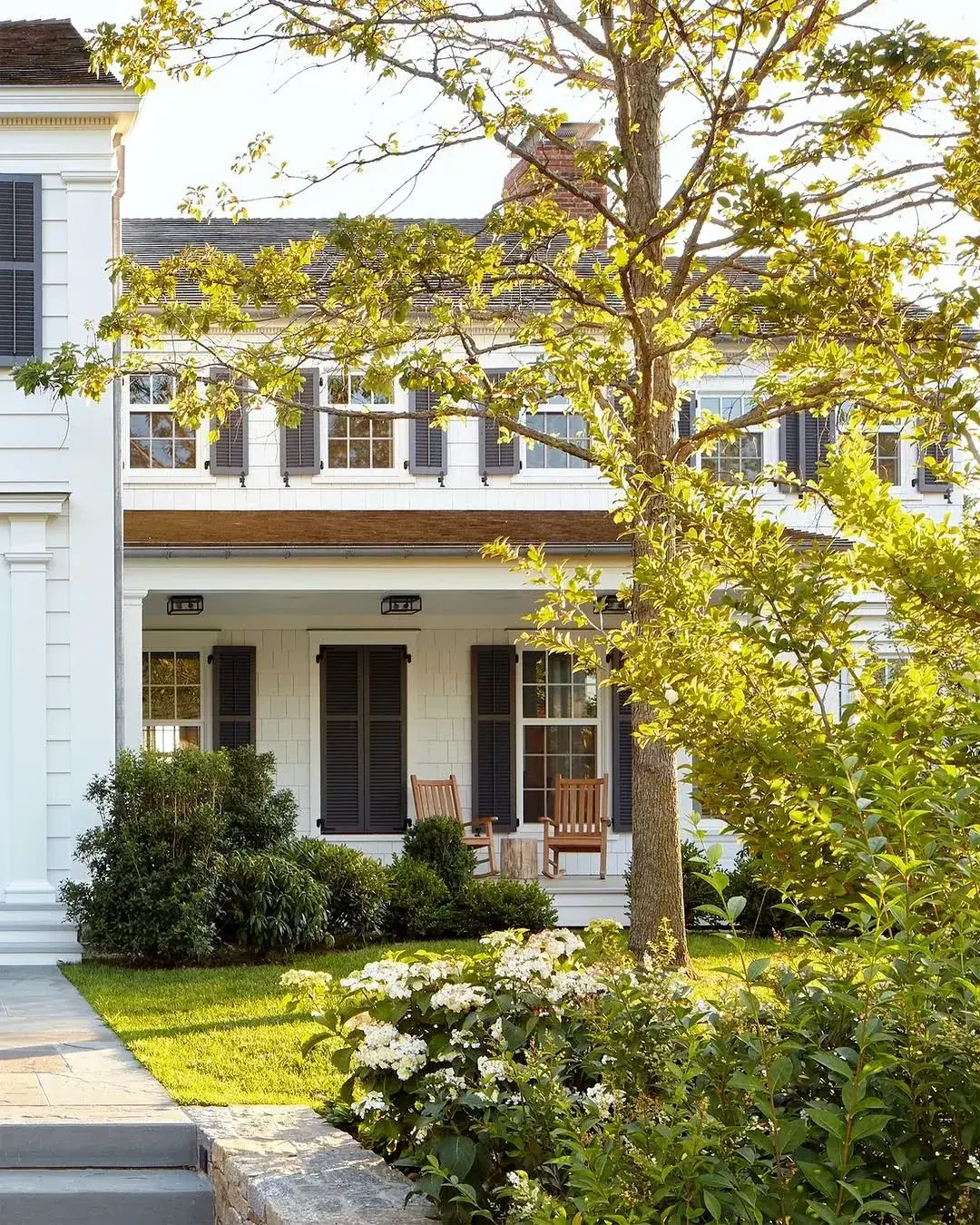 White shingle house with dark louvered shutters on upper windows and on a porch with timber chairs