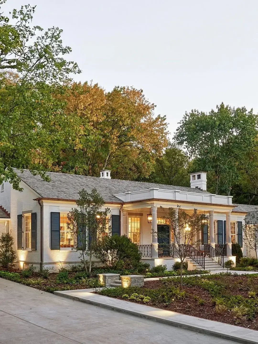 Low white ranch house at dusk with dark shutters on lit windows, cupolas and uplighting