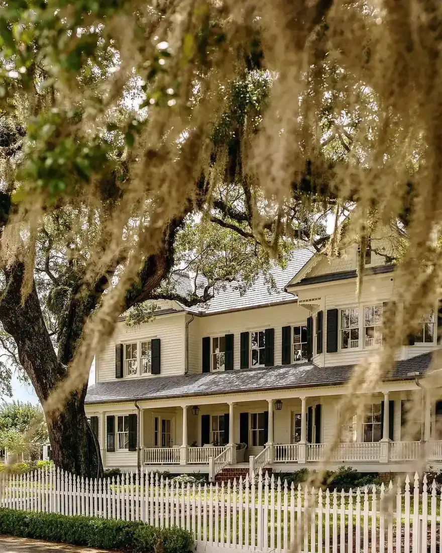 Large cream house with horizontal clapboard, black shutters and a white picket fence