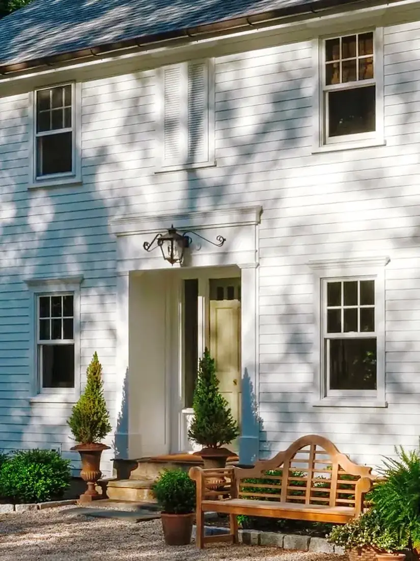 White house with horizontal clapboard, pale blue door, lantern and teak bench