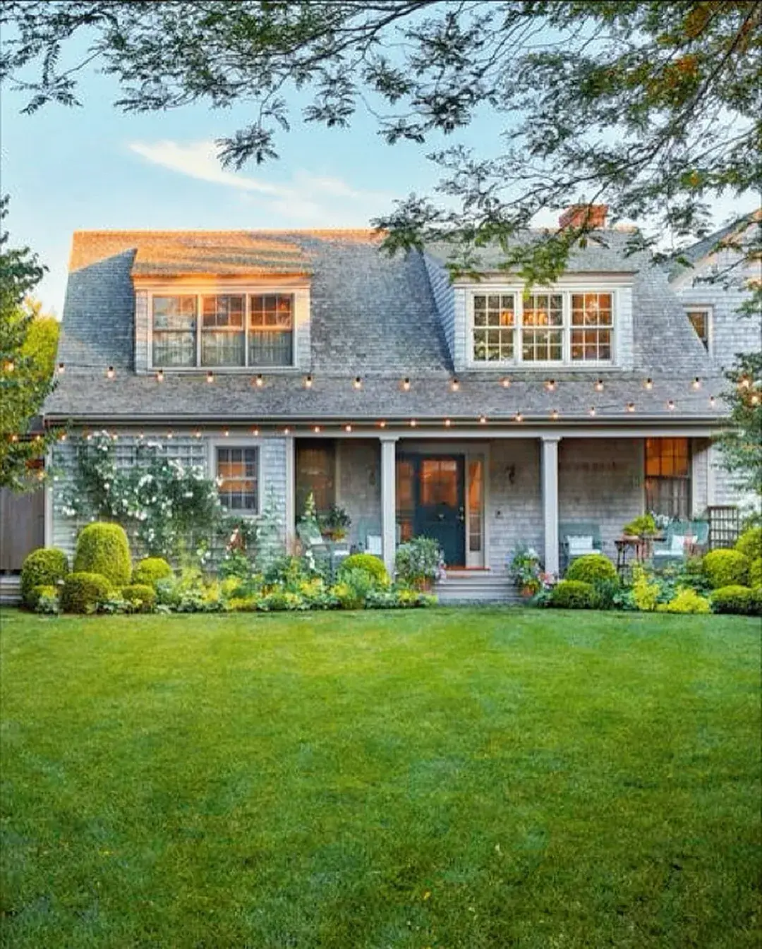 Shingled cottage with string lights hung over a green lawn at dusk