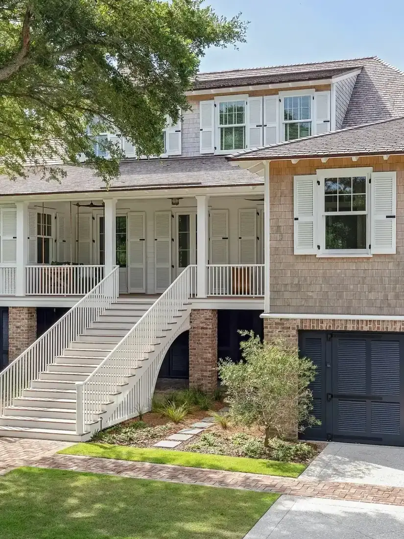 Coastal cottage raised on brick piers with a white double staircase, pale silver shingle above and warmer tan shingle on the lower right wing