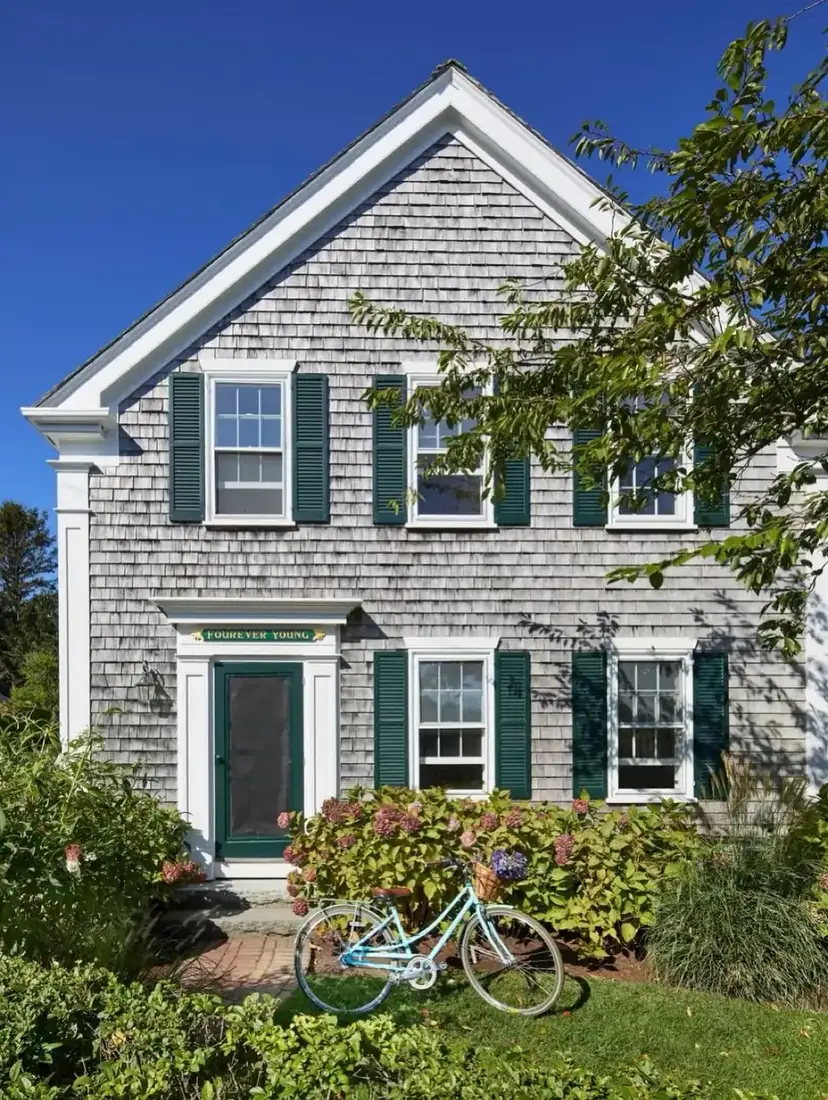 Grey saltbox cottage with silvered cedar walls, white trim, deep green shutters and door, hydrangeas and a pale blue bicycle by the step