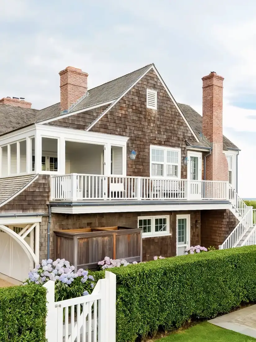 Large coastal house clad in thick brown cedar shakes with white balcony and trim, twin brick chimneys, hydrangeas and a white picket gate