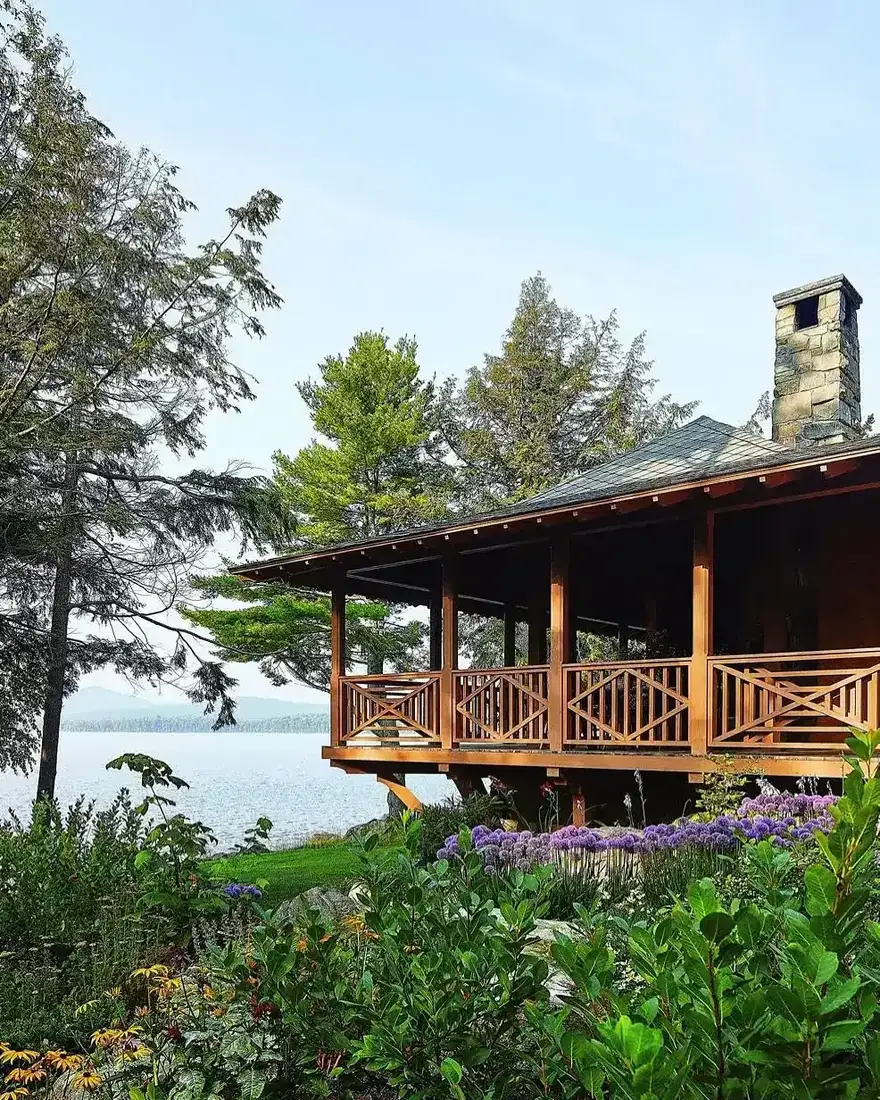 Timber lakeside cottage with a deep porch, cross braced railings, stone chimney, lake and trees beyond, purple alliums and green planting in front
