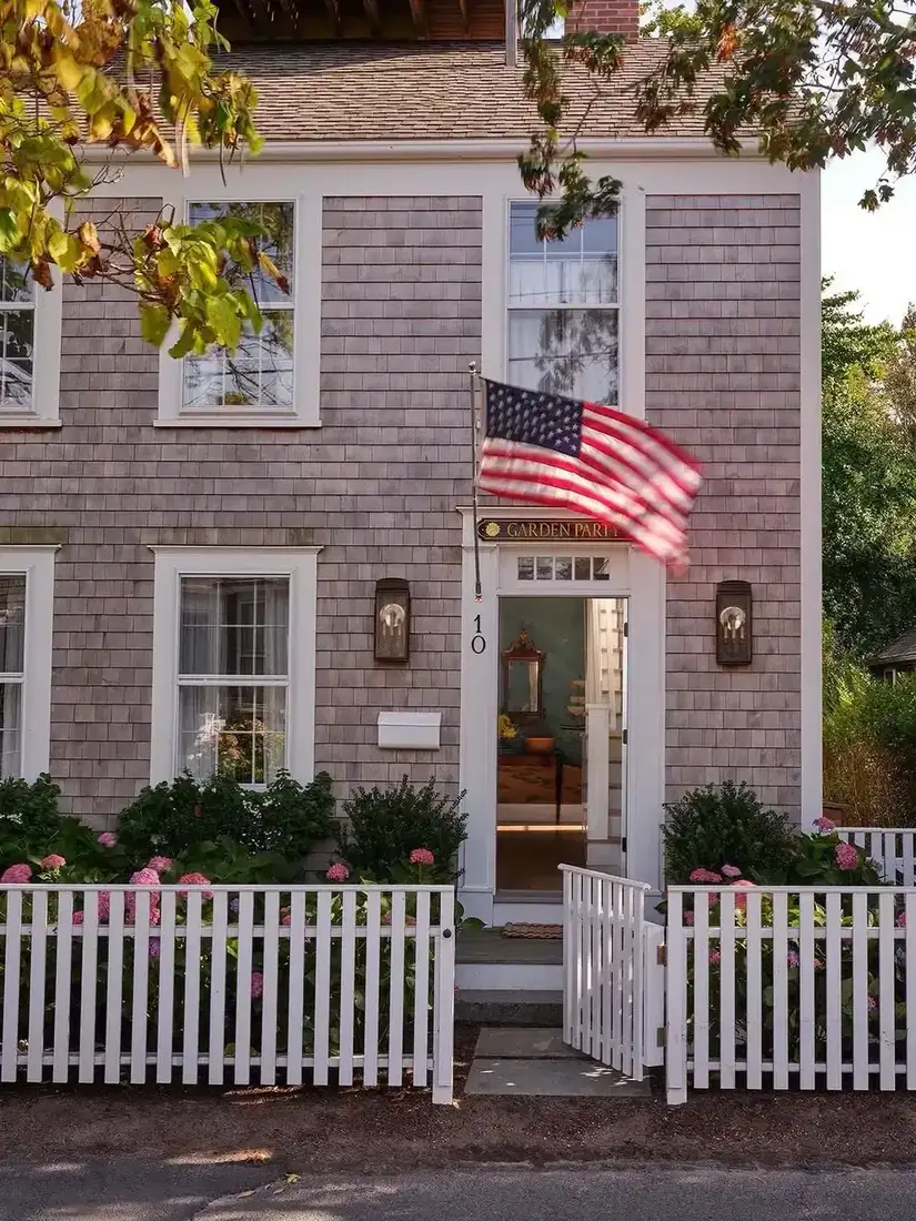 Two storey grey shingle cottage behind a white picket fence and gate, pink hydrangeas, flag over the front door, white trim and warm interior beyond