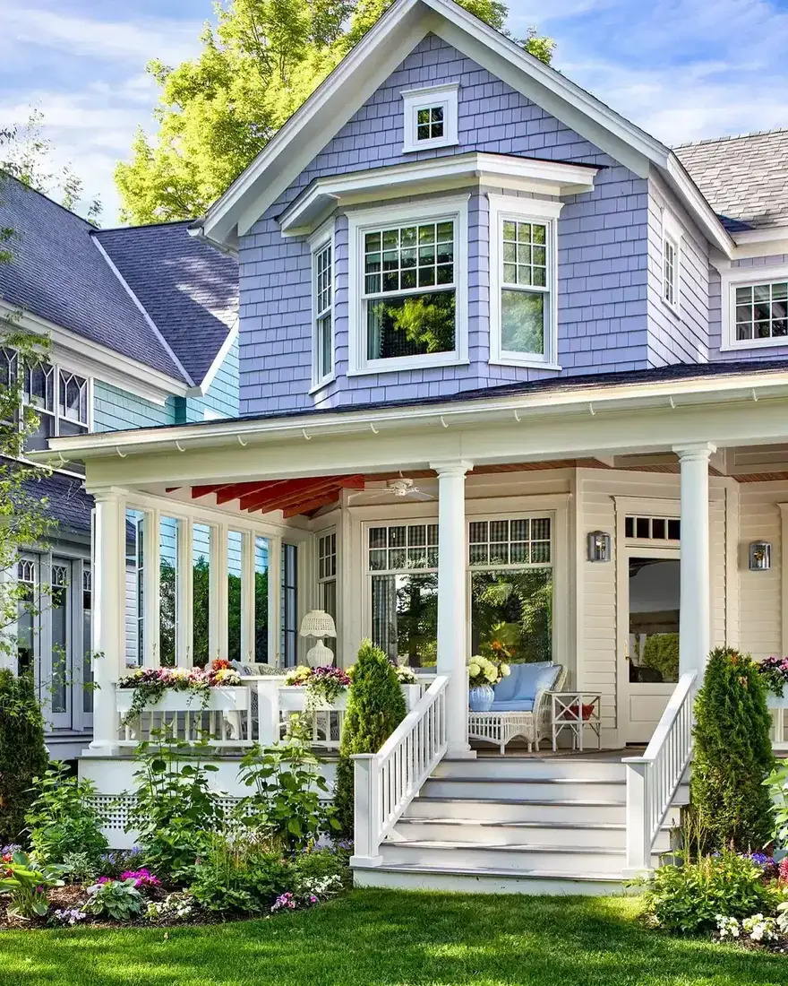Periwinkle blue shingle Victorian house with a deep wraparound porch, bay window and white columns, flower boxes and blue cushions, garden in front