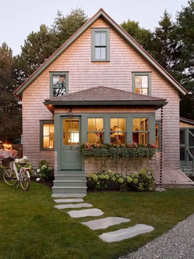 Small gable cottage with pale pinkish natural cedar shingles, sage green door and trim, window box over shingle base, stone slab path and white bicycle