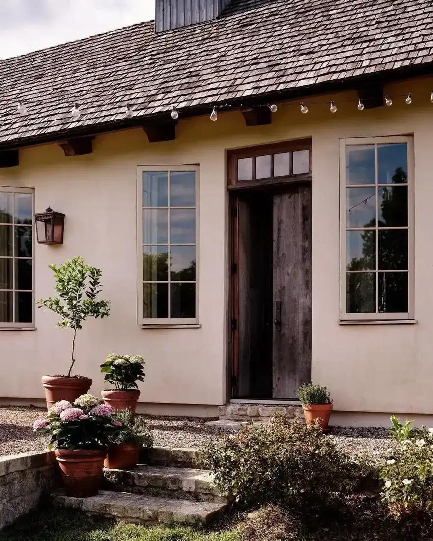 Pale stucco cottage with a silver grey cedar shingle roof, weathered wood door, gravel courtyard and terracotta pots with hydrangeas