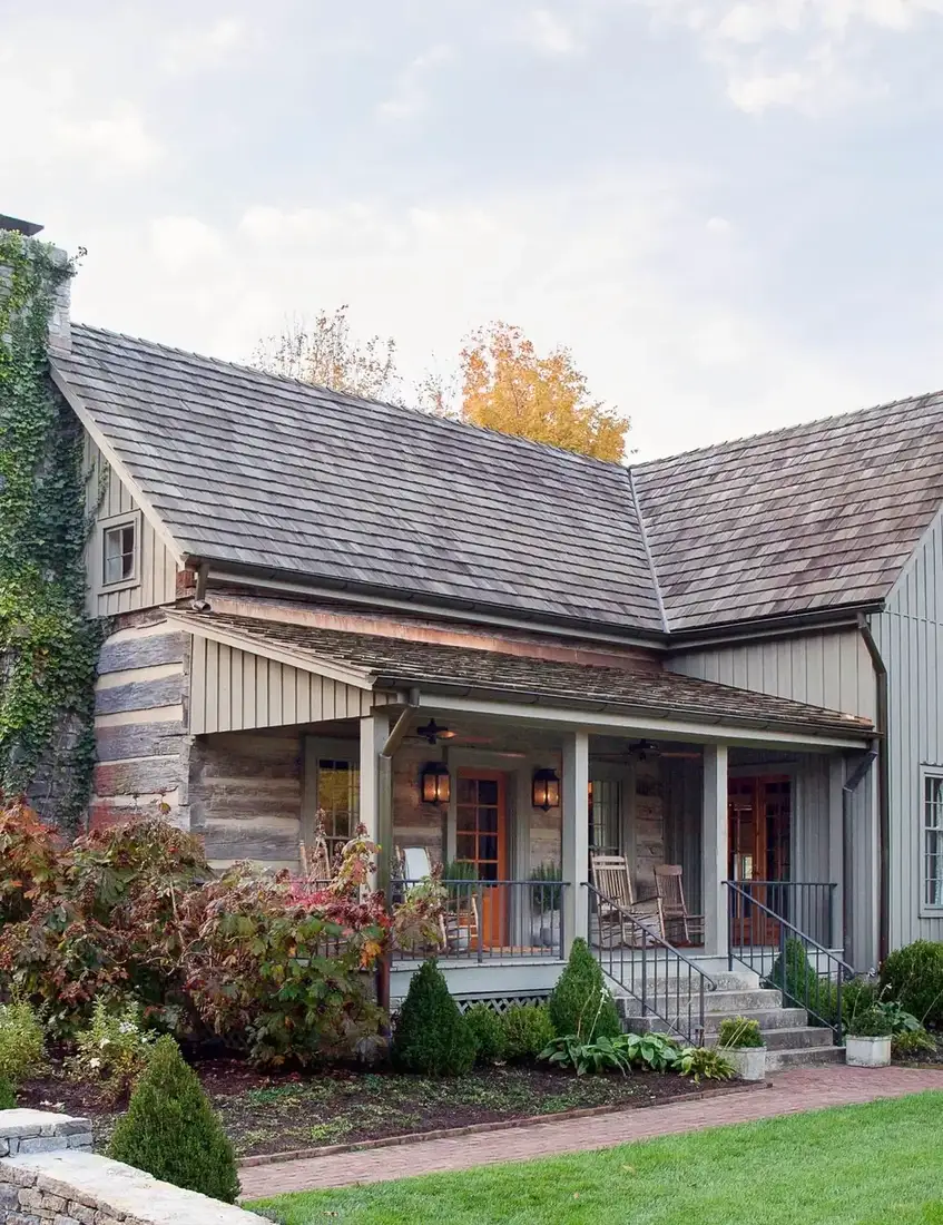 Rustic log and shingle house with a grey cedar roof, stone chimney with ivy, covered porch with rocking chairs and autumn garden