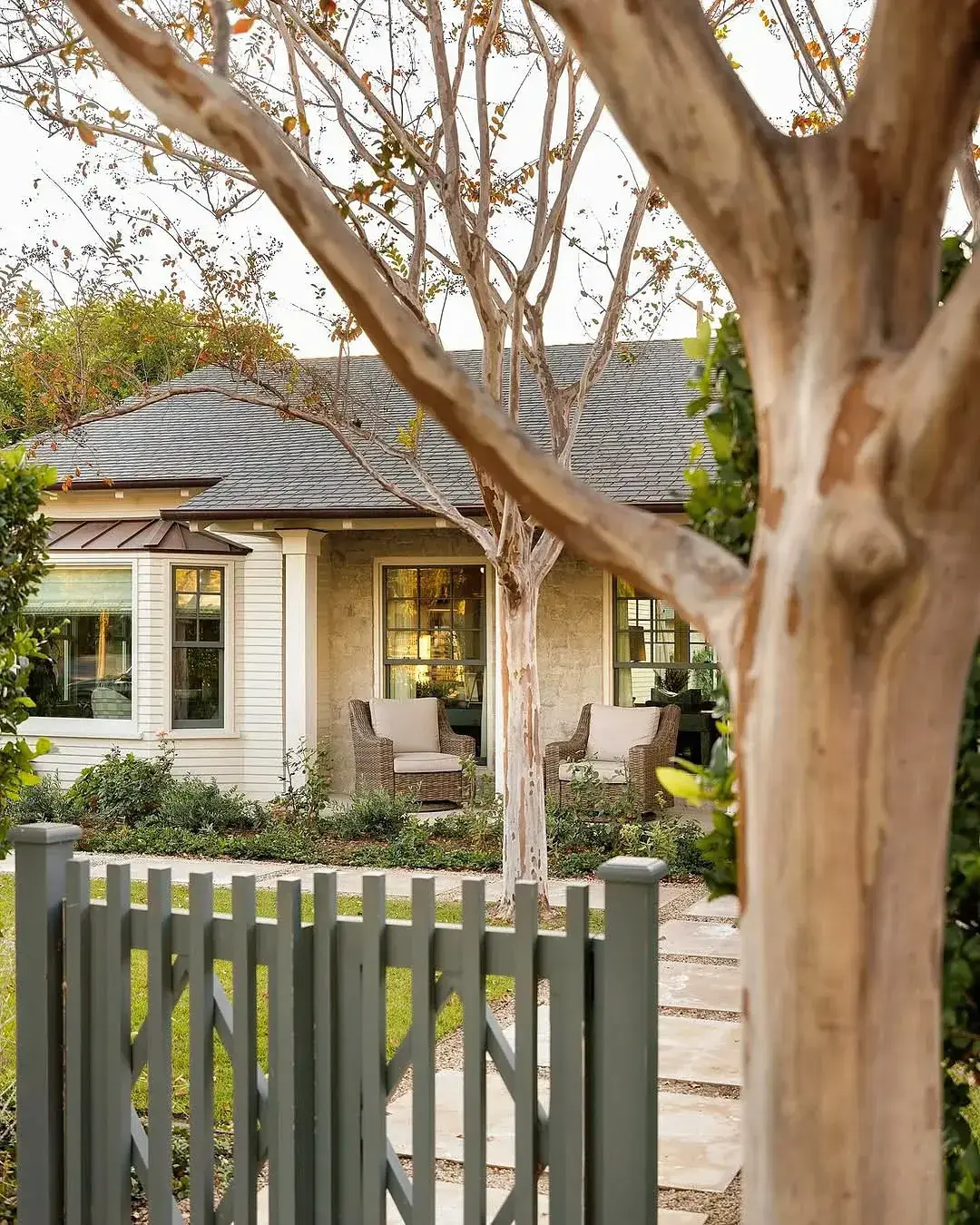 Cream cottage with copper bay roof seen past a grey picket gate and crepe myrtle trunks
