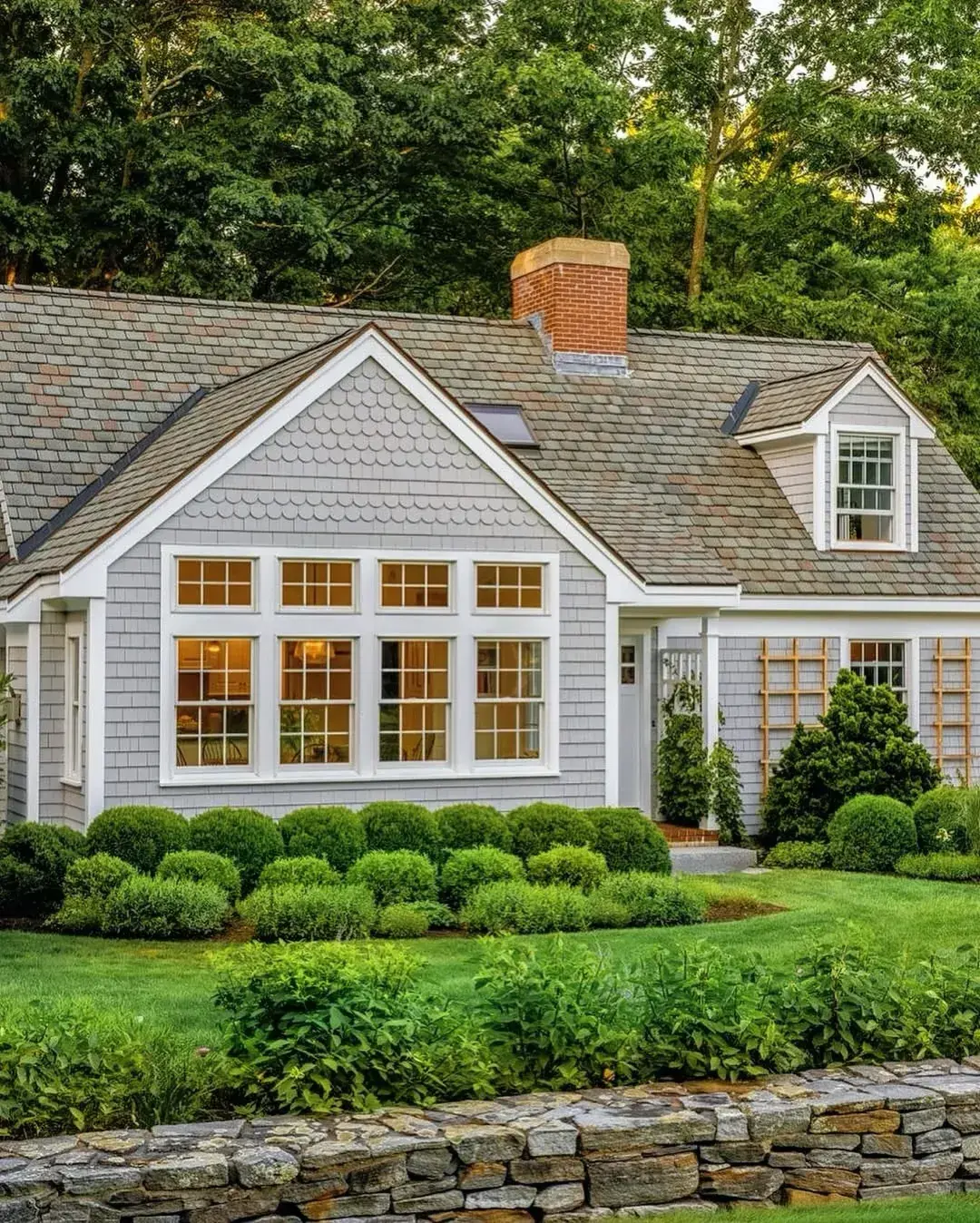 Grey shingle cottage with a wide glazed gable window, clipped box balls and a stone wall