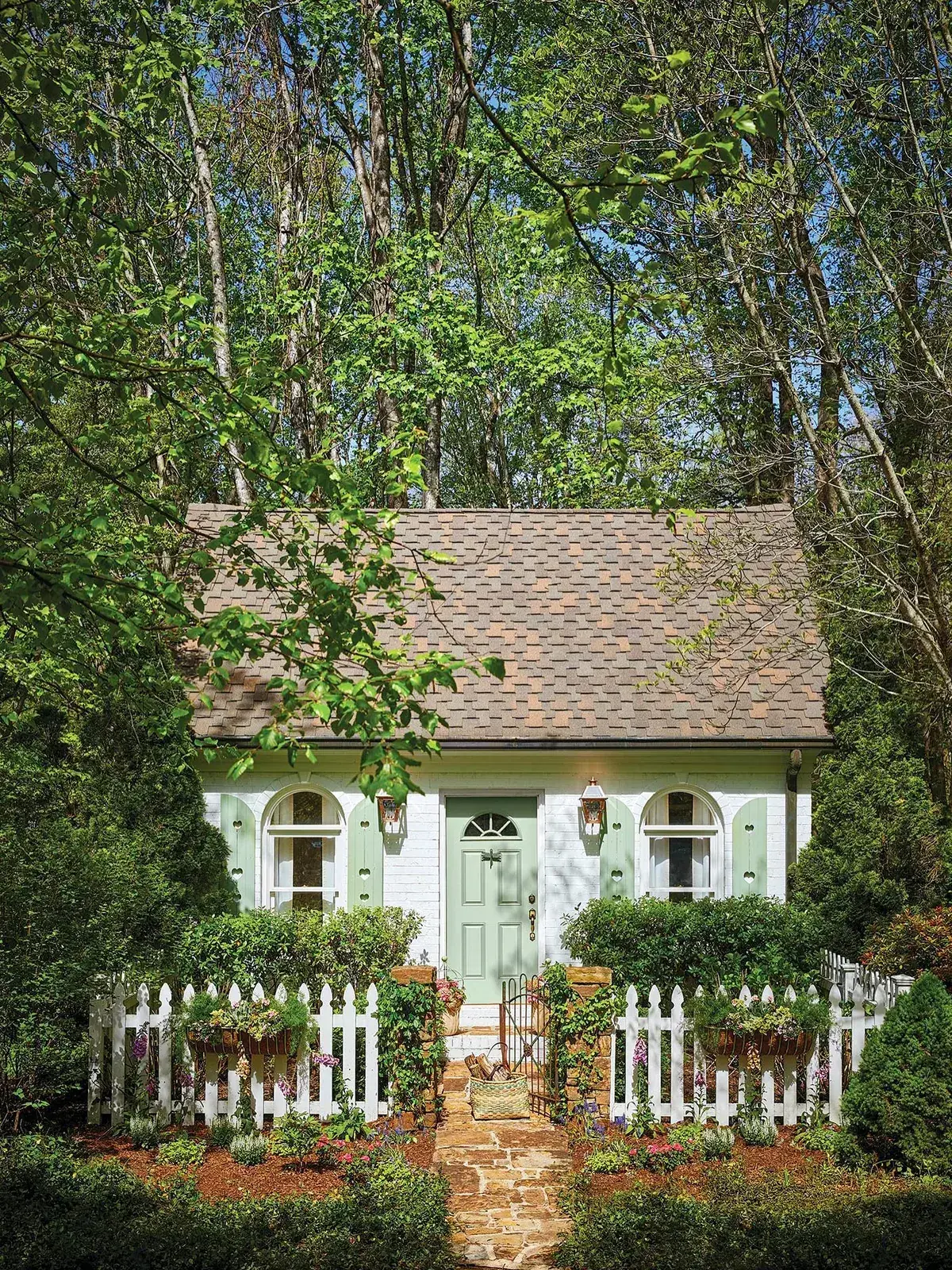 Small white brick cottage with mint door and shutters behind a white picket fence with hanging baskets