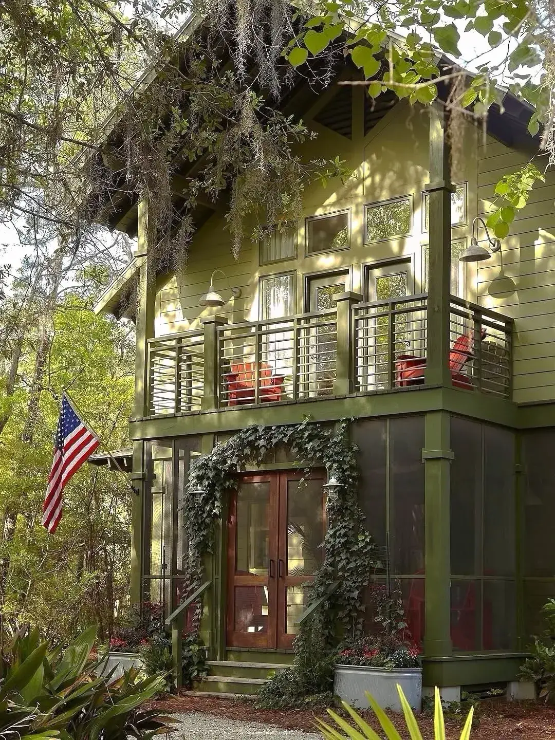 Tall olive green cottage with a first floor balcony, red chairs, flag bracket and ivy clad porch