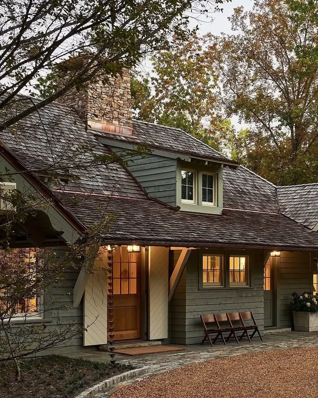 Sage grey cottage at dusk with a low sweeping shingle roof, stone chimney and timber chairs