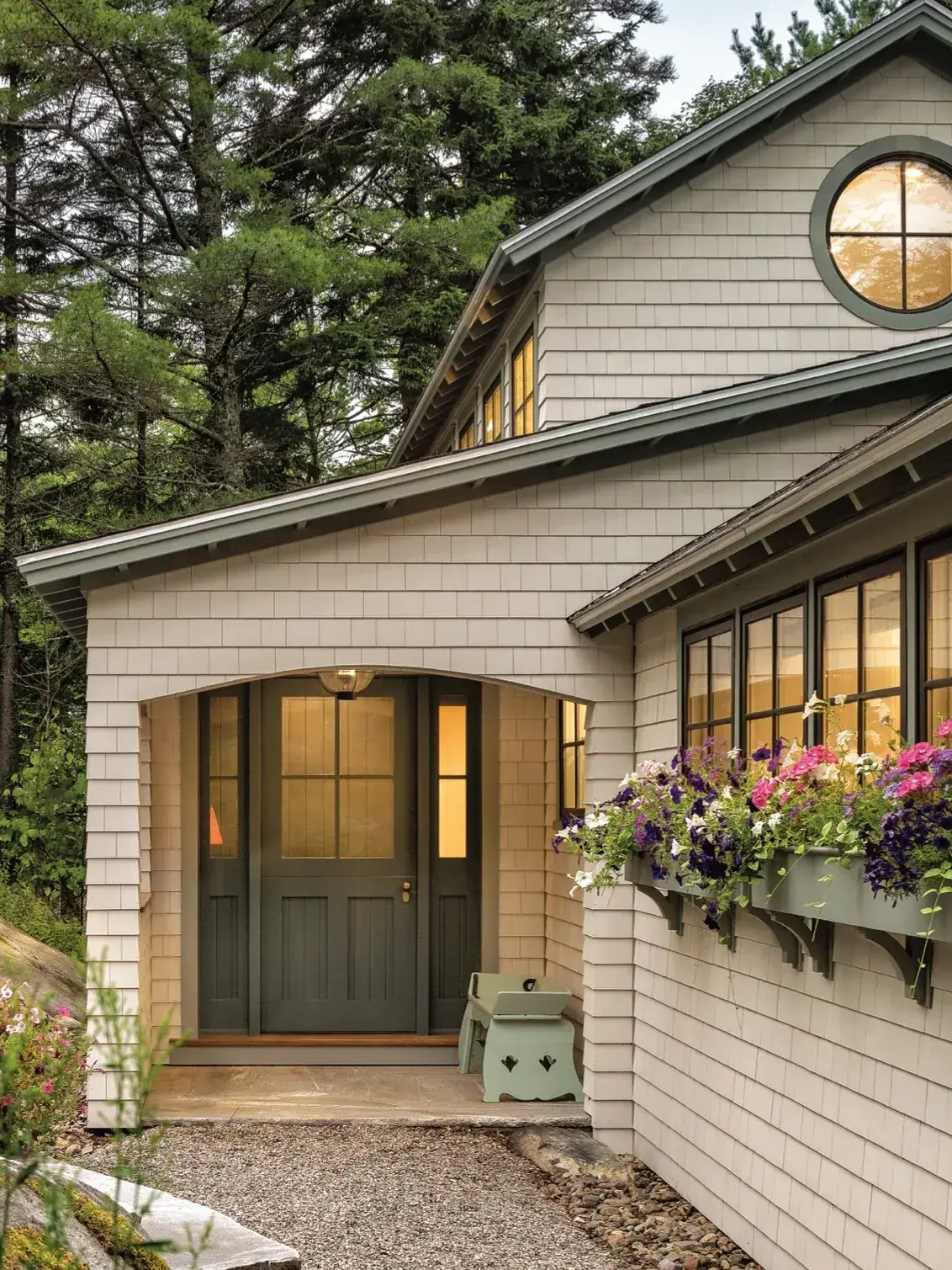 Shingle cottage porch with a sage panelled door, sidelights, round gable window and a flower filled window box