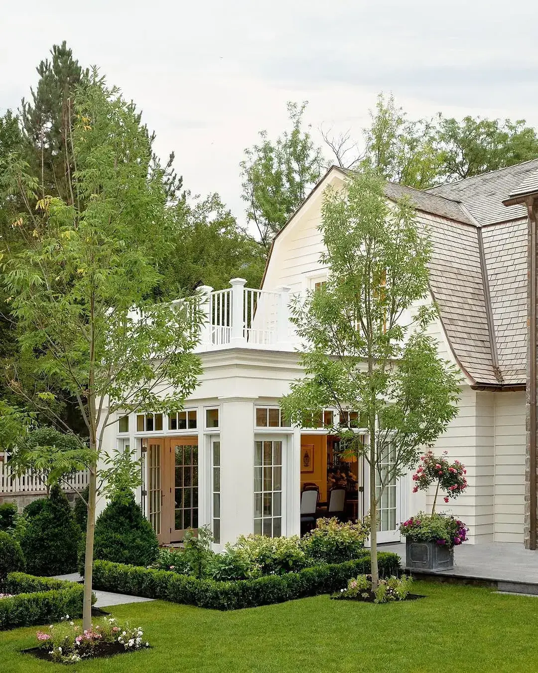 White gambrel cottage with glazed doors, box hedging, young trees and a lead planter