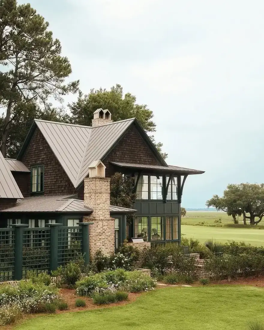 Dark stained shingle cottage with pale metal roof, brick chimney and glazed gable facing a marsh