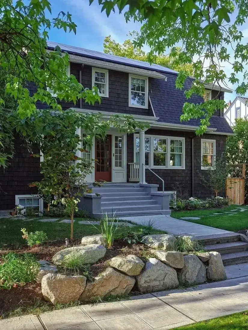 Dark shingle house with white windows behind a boulder retaining wall and young planting