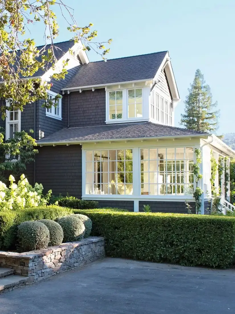 Charcoal shingle house with a white glazed sunroom wing, clipped hedges and stone wall