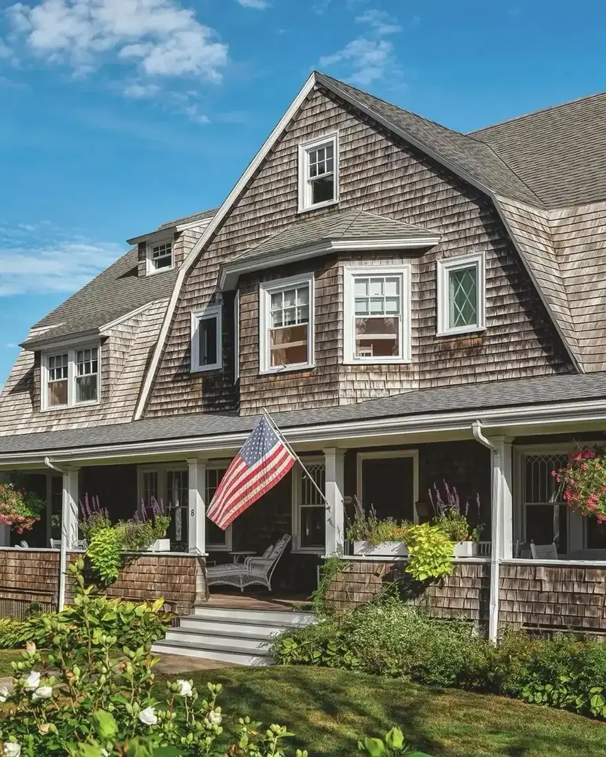 Weathered grey-brown gambrel with a shaded porch, white columns and a flag